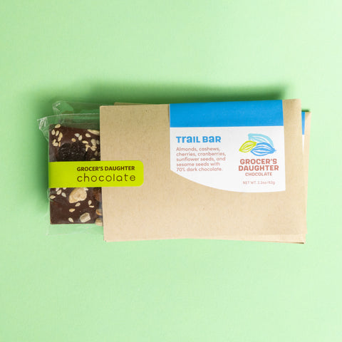 Packaged trail bar with visible dark chocolate pieces and seeds through the transparent wrapping, branded as 'Grocer's Daughter Chocolate'.